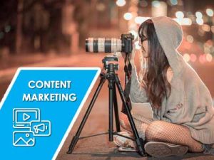 A smiling girl sitting crossed legged looking through a camera secured on a tripod outside at night.